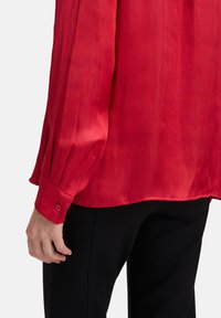 Blouse en satin rouge avec une texture lisse, dotée de manches longues et de poignets boutonnés, associée à un pantalon noir.