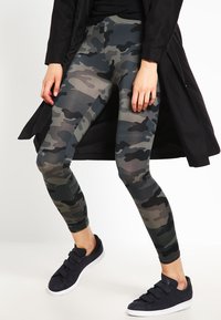 Leggings u kamuflaži u nijansama zelene i sive, s pripijenim dizajnom. Uparene s crnim tenisicama na zatvaranje i dugim crnim kaputom.