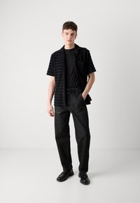 Svartstripete kortermet skjorte over en svart T-skjorte, kombinert med løse svarte jeans og svarte sko; har en knapp-elastisk design.