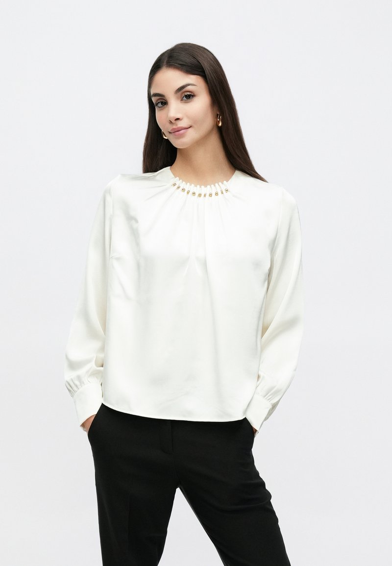 Hvid satinbluse med lange ærmer, rynket halsudskæring og dekorative gyldne detaljer. Kombineret med sorte bukser, hvilket skaber et kontrastfuldt look.