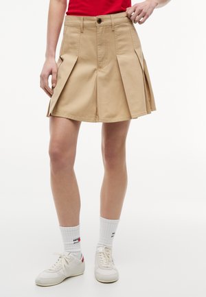 Vrouw draagt een beige geplooide rok, witte sokken met logo en witte sneakers, en past de tailleband van de rok aan.