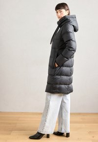 Abrigo de plumas negro con cuello alto y capucha, que presenta un diseño acolchado y bolsillos laterales. Combinado con unos vaqueros de pierna ancha de color azul claro y botas negras.