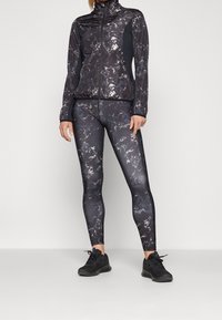 Černá sportovní bunda s vysokým límcem, zdobená mramorovým vzorem. Černé legíny se shodným designem a jednobarevnými černými akcenty. Černé tenisky.