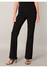 Frau trägt enge schwarze Hose und schwarze Sandalen mit dünnen Riemen und hohen Absätzen, steht vor einem neutralen Hintergrund.