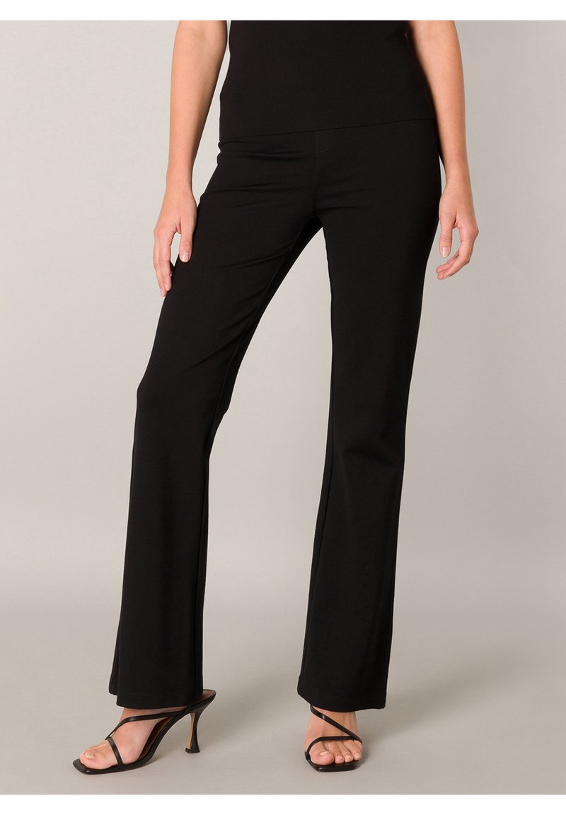 Frau trägt enge schwarze Hose und schwarze Sandalen mit dünnen Riemen und hohen Absätzen, steht vor einem neutralen Hintergrund.