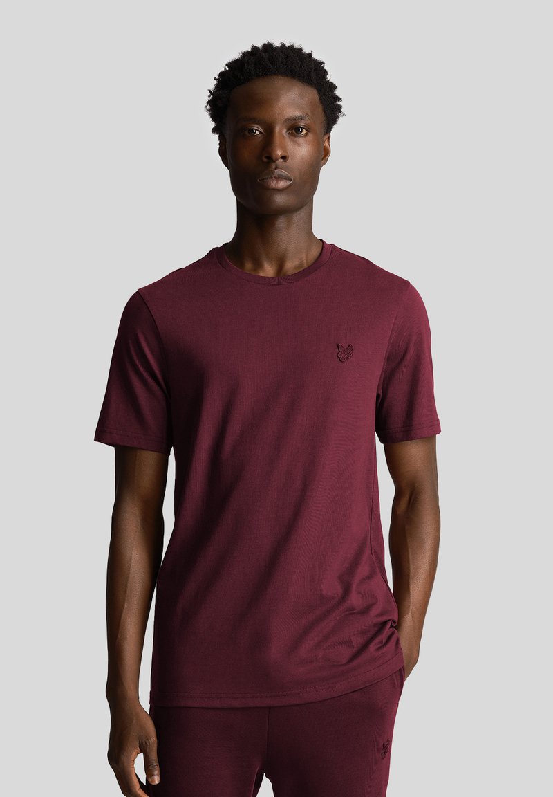 T-shirt di cotone bordeaux con maniche corte, scollatura rotonda e un piccolo logo sul lato sinistro del petto. Texture liscia, design casual.