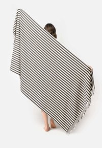 La coperta a righe presenta linee alternate marroni e bianche, con bordi frangiati. Il tessuto leggero si drappeggia elegantemente, sostenuto da una persona.