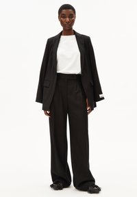 Schwarze taillierte Blazer über einem weißen Hemd, kombiniert mit weit geschnittenen schwarzen Hosen. Auffällige Details sind subtile Akzente an den Blazerärmeln.