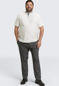 Man standing with hands in pockets wearing a white short-sleeve henley shirt, gray pants, and black shoes against a plain background.