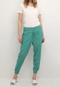 Tealfärgade mönstrade joggerbyxor med elastisk midja och manschetter vid anklarna, kombinerat med en vit V-ringad t-shirt och vita snörningssandaler.