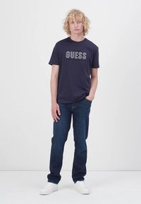 Navy short-sleeve t-shirt with "GUESS" logo, paired with dark blue jeans and white sneakers. Model stands against a plain background.