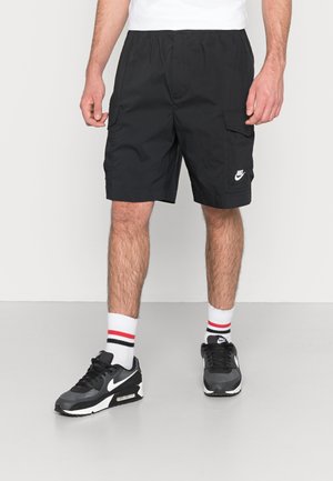 Mężczyzna ubrany w czarne szorty cargo Nike, białe skarpety za kostkę z czerwono-czarnymi paskami oraz czarne buty sportowe Nike stoi na szarej podłodze.