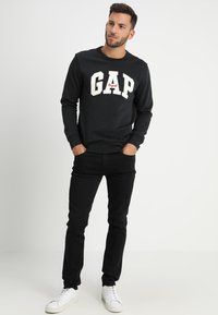 Black GAP sweatshirt with white lettering and red accent, paired with black jeans and white sneakers. Model stands with hands in pockets.