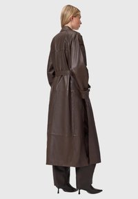 Brązowy skórzany płaszcz trencz z paskiem w talii, długimi rękawami i bocznymi rozcięciami. Charakteryzuje się gładką fakturą i minimalistycznym designem.
