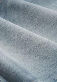 Svijetloplavi denim materijal s teksturiranim tkanjem, vidljivim šavovima i blago izblijedjelim izgledom. Općenito glatka površina.