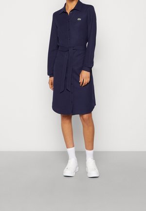 Vrouw draagt een marineblauwe blousejurk met knopen en een tailleceintuur, witte sokken en witte sneakers, staand tegen een effen witte muur.
