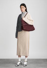 Μπορντώ σουέντ τσάντα χεριού με σχέδιο που διπλώνει, κρατημένη από μοντέλο που φοράει γκρι πουλόβερ με πλέξη καλωδίων και μπεζ midi φούστα, συνδυασμένη με μαύρες ψηλοτάκουνες σανδάλια.