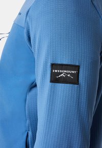 Blå langærmet udendørsjakke med tekstureret stof og sort rektangulært mærke på overarmen med teksten "Swedemount" og bjerglogo.