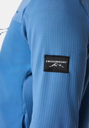 Blå stofjakkeærme med et struktureret mønster, en sort rektangulær lap med "SWEDEMOUNT" og et bjerggrafik.
