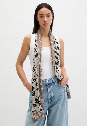 Kobieta o prostych ciemnych włosach nosi biały top bez rękawów, niebieskie jeansy z wysokim stanem oraz długi beżowy szalik z czarnym kwiatowym wzorem, ręce w kieszeniach.