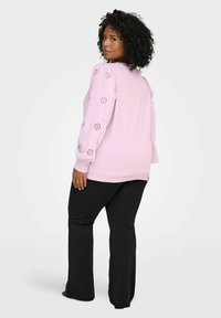 Svetloroza pulover z napihnjenimi rokavi in rezanimi cvetličnimi vzorci, kombiniran s črnimi razširjenimi hlačami, pogled od zadaj.