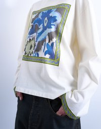 Άτομο που φοράει κρεμ πουκάμισο μακρυμάνικο με μπλε φλοράλ σχεδιασμό και πράσινο περίγραμμα, με τα χέρια στις τσέπες, σε συνδυασμό με σκούρο τζιν παντελόνι.