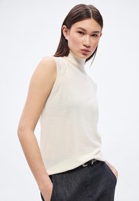 Ujjatlan, krémszínű felső magas nyakkal, puha anyagból készült. Testhezálló szabású, finom textúrával, sötét nadrággal párosítva.