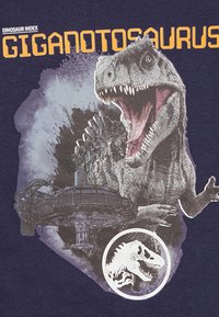 Marinefarbendes T-Shirt mit einem Grafikdesign eines Giganotosaurus in Grautönen, begleitet von auffälligem orangefarbenem Text und einem detaillierten, runden Dinosaurier-Logo.