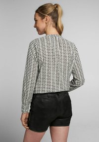 Schwarz-weiße karierte Bluse mit langen Ärmeln und gerafften Details, kombiniert mit taillierten schwarzen Shorts. Der Stoff wirkt leicht und strukturiert.