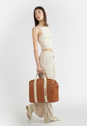 Giovane donna con top beige senza maniche e pantaloni larghi che porta una grande borsa a tracolla marrone con manici crema, guardando oltre la spalla.