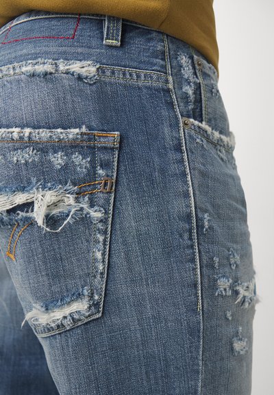 Vaqueros de mezclilla azul desgastados con bordes deshilachados y múltiples detalles rasgados. Cuenta con un diseño tradicional de cinco bolsillos y costuras visibles.