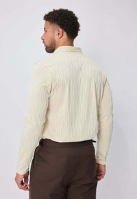 Chemise à manches longues couleur crème avec un motif texturé, col classique, et silhouette droite, associée à un pantalon marron.