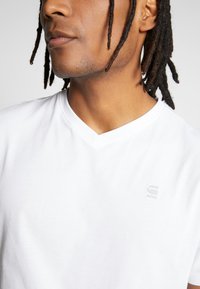 Weißes Baumwoll-T-Shirt mit V-Ausschnitt und einem kleinen, dezenten Logo auf der Brust. Glatte Textur mit kurzen Ärmeln.