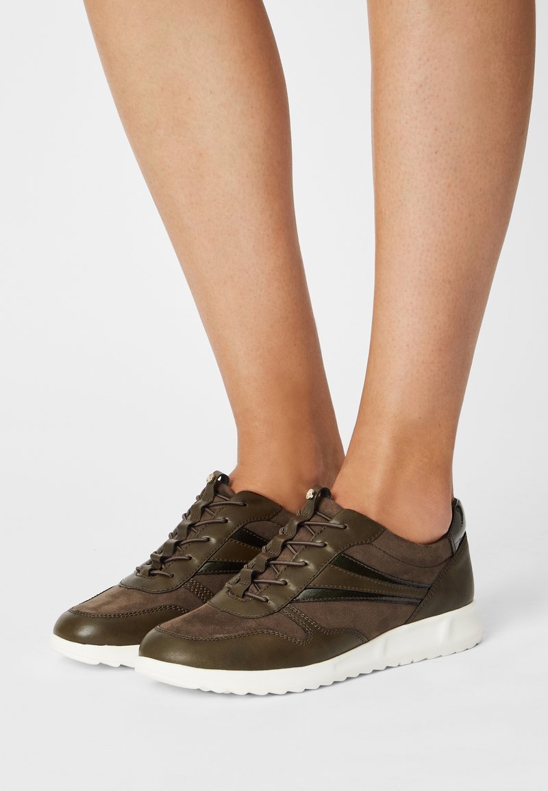 Jambes portant des baskets en cuir et en daim marron avec des bandes noires et des semelles blanches, posées sur une surface blanche.