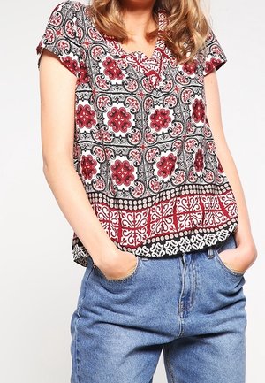 Vrouw draagt een kortermige blouse met rood-zwarte patronen, ingestopt in hooggetailleerde blauwe jeans, handen in de zakken.