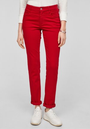 Vrouw met felrode opgerolde broek, witte sneakers en een witte top met lange mouwen, staand voor een effen achtergrond.