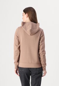 Sudadera con capucha marrón de textura suave, con mangas ajustadas y dobladillo acanalado. Llevada con pantalones gris oscuro, vista desde atrás.