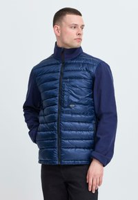 Mornarska puffer jopica z zadrgo spredaj, horizontalnim kvačkanim vzorcem, visokim ovratnikom in kontrastnimi modrimi rokavi. Sprednji žep in slim fit oblikovanje.