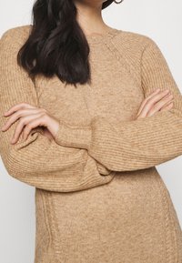 Beige gestrickter Pullover mit strukturierten Rippenbündchen, runder Ausschnitt und sichtbaren Nähten an den Seiten. Bequem und lässige Passform.