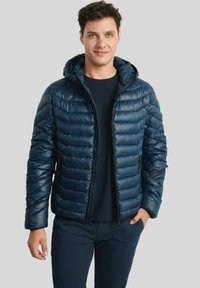 Mężczyzna ubrany w granatową pikowaną kurtkę z kapturem, noszącą ciemną koszulę, z rękami w luźnej pozycji, stojący na jednolitym szarym tle.