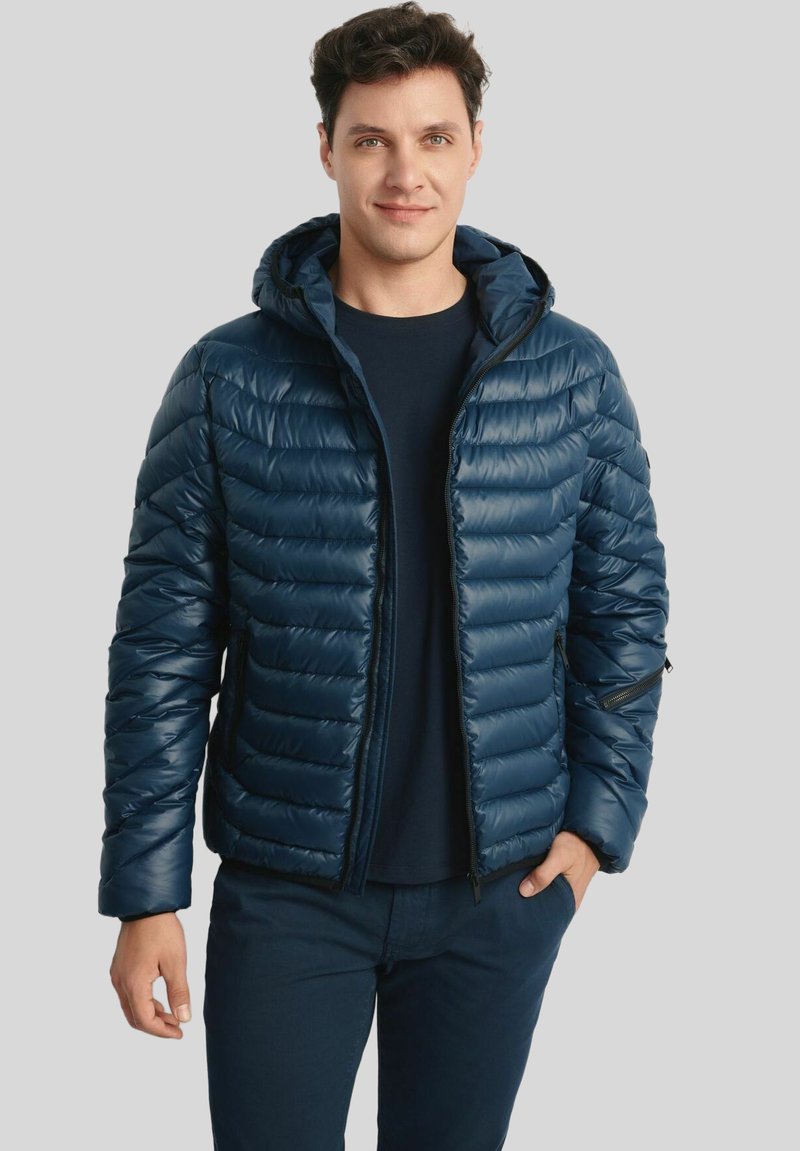 Mężczyzna ubrany w granatową pikowaną kurtkę z kapturem, noszącą ciemną koszulę, z rękami w luźnej pozycji, stojący na jednolitym szarym tle.