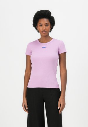 Lilafarbenes, kurzärmliges, eng anliegendes T-Shirt aus Rippstoff mit rundem Ausschnitt und einem kleinen blauen Etikett auf der Vorderseite. Kombiniert mit schwarzen Hosen.