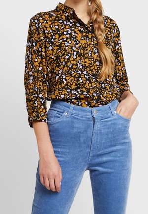 Frau trägt eine langärmelige schwarze Bluse mit orangefarbenen und violetten abstrakten Flecken, kombiniert mit hochtaillierten blauen Cordhosen.