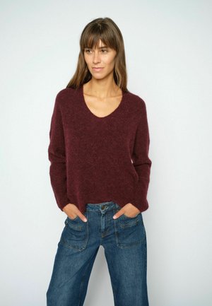 Donna con capelli castani lisci che indossa un maglione bordeaux con scollo a V e jeans blu in denim con le mani nelle tasche davanti a uno sfondo semplice.