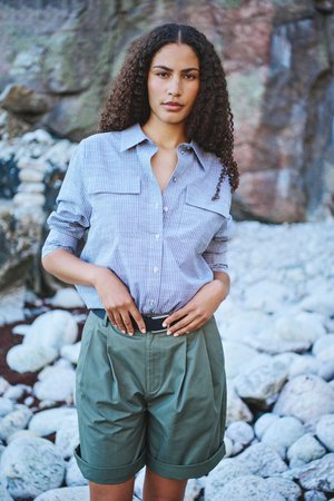 Naisel lokkis juustega, kandes triibulist nööpidega särki ja rohelist värvi mansettidega lühikesi pükse, seisab kivisel maastikul kaljuse taustaga.