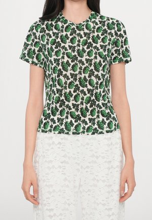 Kobieta w białym topie z krótkim rękawem z zielonym wzorem roślin chmielu i białych spodniach z koronkowym wzorem kwiatowym, stojąca na jednolitym tle.