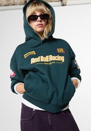 Blond persoon met een donker teal Red Bull Racing hoodie en zwarte zonnebril staat met de handen in de zakken tegen een neutrale achtergrond.