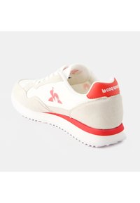 Scarpa da ginnastica bianca con dettagli in camoscio, marchio rosso e suola testurizzata. Il design presenta una punta arrotondata e un colletto imbottito.