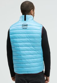 Hellblaues Puffer-Westen mit steppendem Design und hohem Kragen, mit schwarzem Reißverschluss und bedrucktem Text auf der Rückseite. Darunter ein schwarzes Shirt.