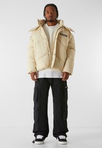 Beige padded jacket with diagonal stitching and a chest pocket. Worn over a cream sweater and paired with black cargo pants and sneakers.